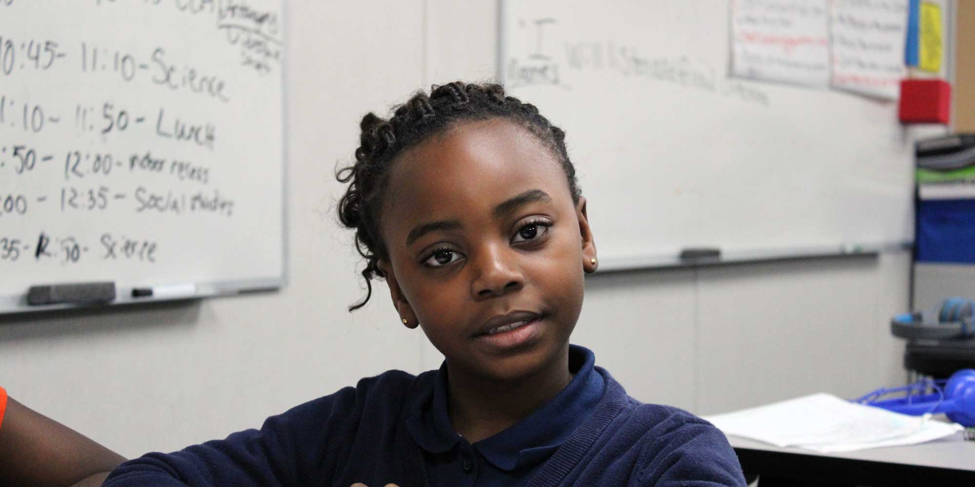 Student seated in a classroom in front of a whiteboard class schedule.